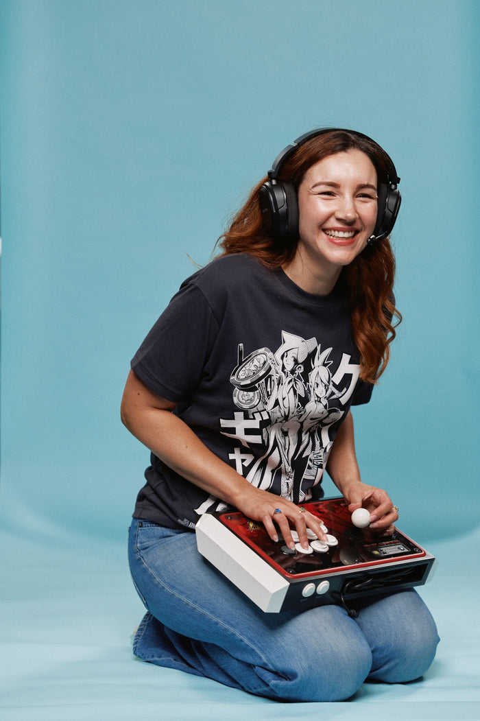 A woman kneeling on a light blue studio backdrop, wearing over-ear headphones and a black graphic T-shirt while using a red and white arcade controller on her lap.