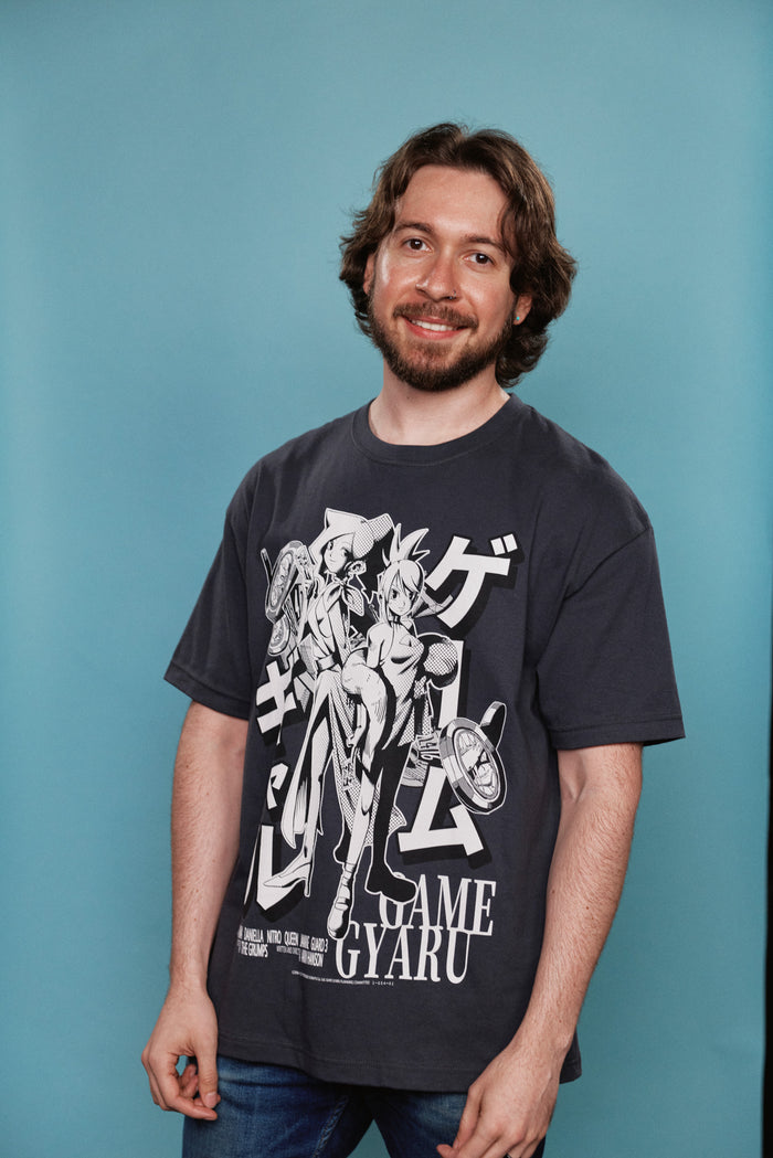 A man with brown hair and a beard wearing a dark gray graphic t-shirt with a white anime print, standing against a solid teal-blue background.