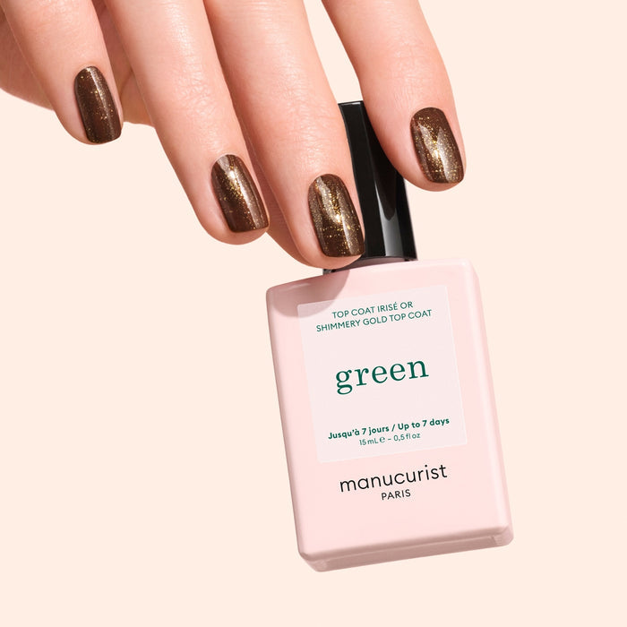A hand with short, shimmery dark brown nails holds a bottle of Manucurist Glitter Top Coat, featuring a gel-like finish, against a light peach background.