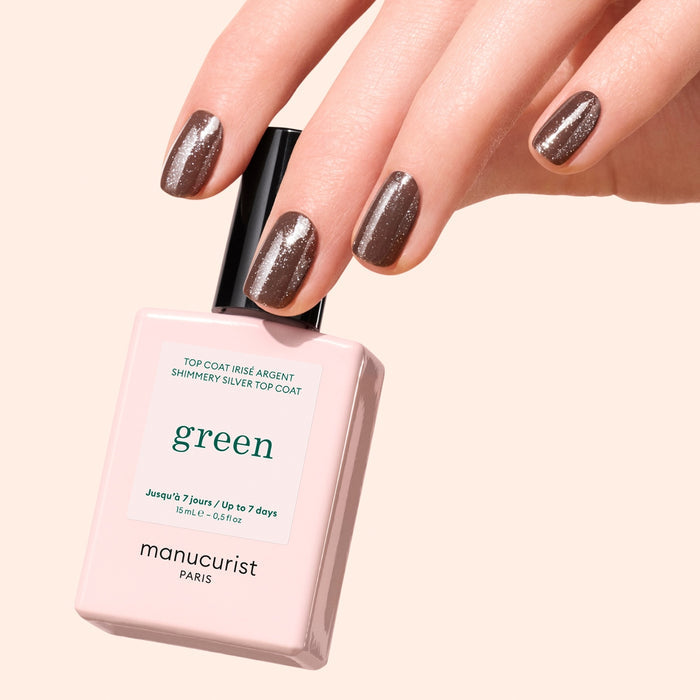 A hand with manicured nails painted in shimmery brown polish holds a bottle of Manucurist Glitter Top Coat, highlighting a dazzling gel-like finish against a light beige background.
