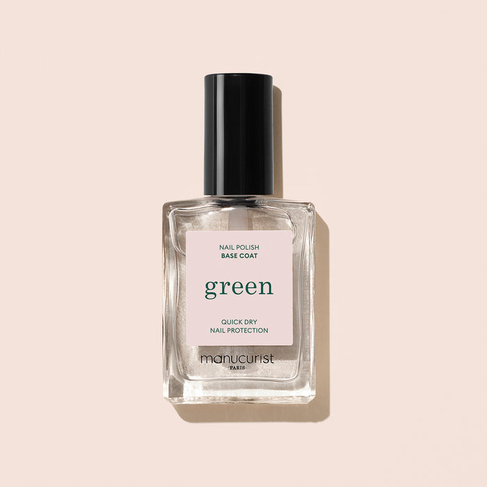 A clear glass bottle of Manucurist Green Base Coat with a black cap sits against a beige background. The label highlights its quick-dry, nail protection formula—ideal for prolonging polish wear and effective as a treatment.