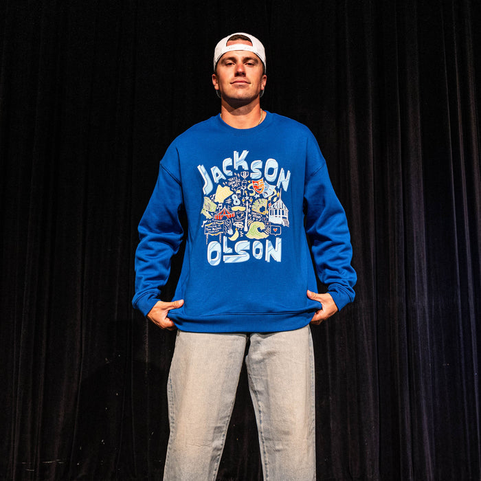Adult male model on a stage wearing the royal blue Jackson Olson x BL101 Hometown Crewneck showing the full front "Jackson Olson" graphic with colorful illustrated artwork