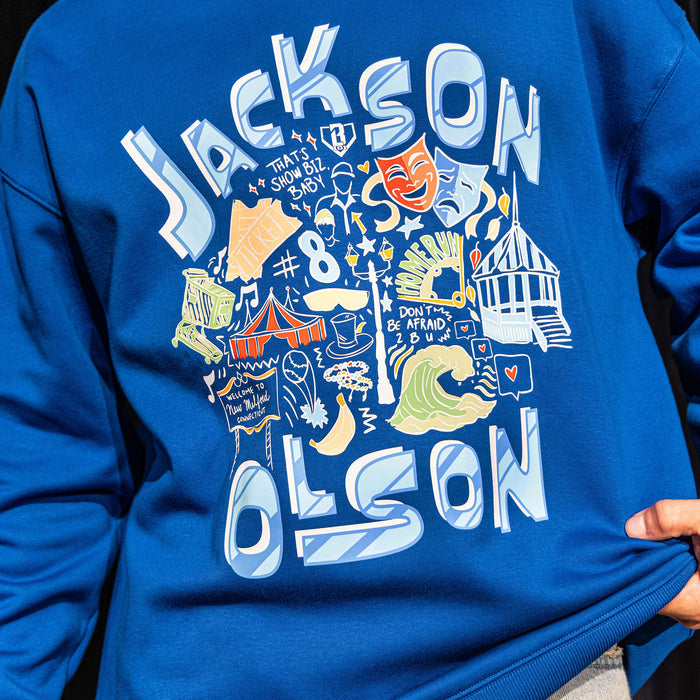 Close-up of the front graphic on the royal blue Hometown Crewneck, showing the "Jackson Olson" lettering surrounding colorful illustrated artwork including theater masks, waves, circus tent, and "That's Show Biz, Baby" text