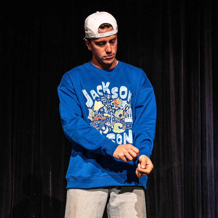 Adult male model adjusting his sleeve on a stage wearing the royal blue Jackson Olson x BL101 Hometown Crewneck with jeans