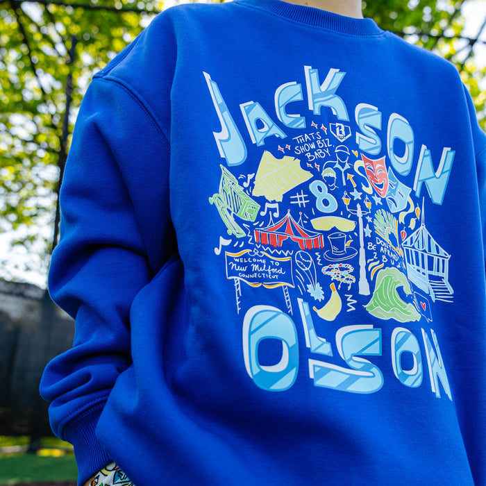 Smiling youth player standing on a baseball field wearing the royal blue Jackson Olson x BL101 Hometown Crewneck paired with the white Hometown Shorts