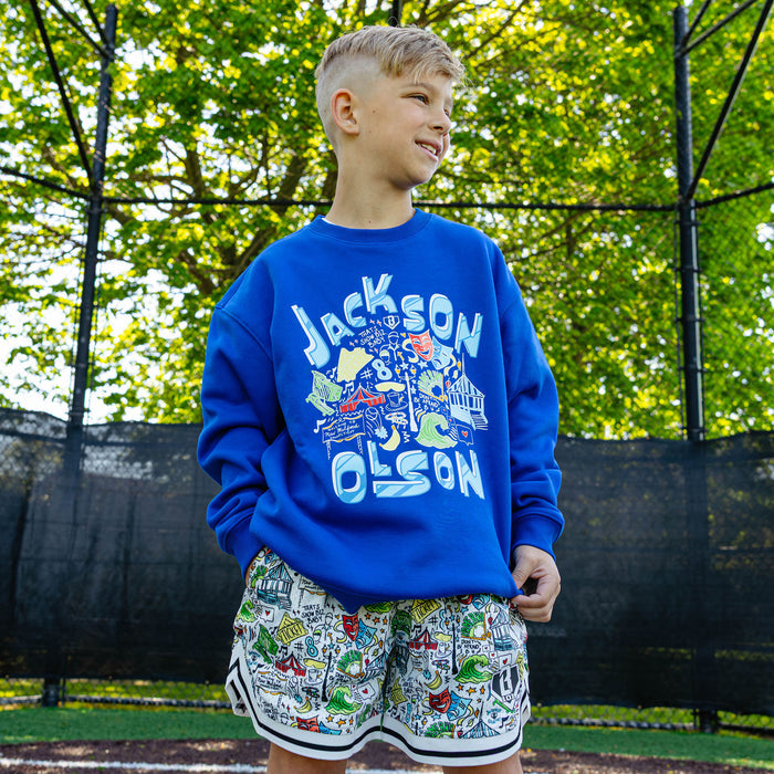 Close-up of the front graphic on the youth royal blue Hometown Crewneck showing the "Jackson Olson" lettering surrounding colorful illustrated artwork including theater masks, circus tent, and "That's Show Biz, Baby" text