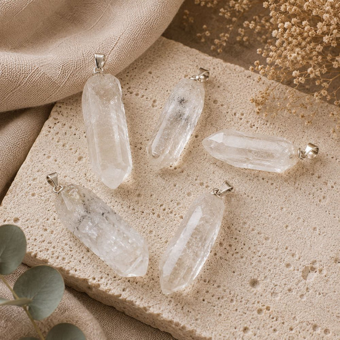 Five clear quartz crystal point pendants with silver bail loops, arranged on a textured beige stone slab with neutral fabric and dried floral props.