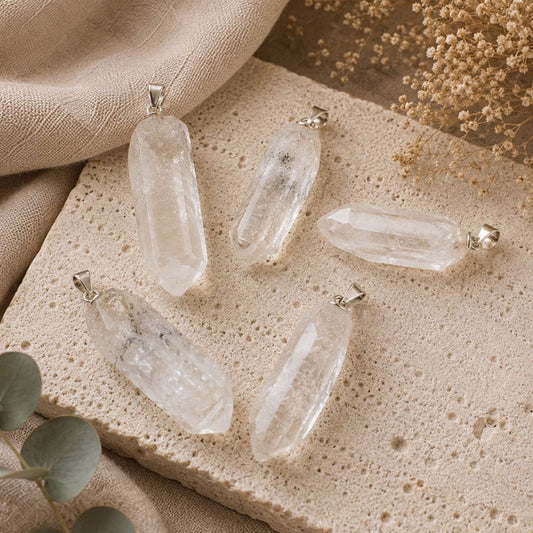 Five clear quartz crystal point pendants with silver bail loops, arranged on a textured beige stone slab with neutral fabric and dried floral props.