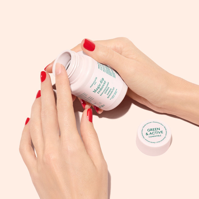 A person with red-painted nails opens a white Magic Dip Remover bottle by Manucurist, labeled “Magic Dip Remover,” against a light beige background. The travel-friendly cap with the Manucurist branding lies nearby.