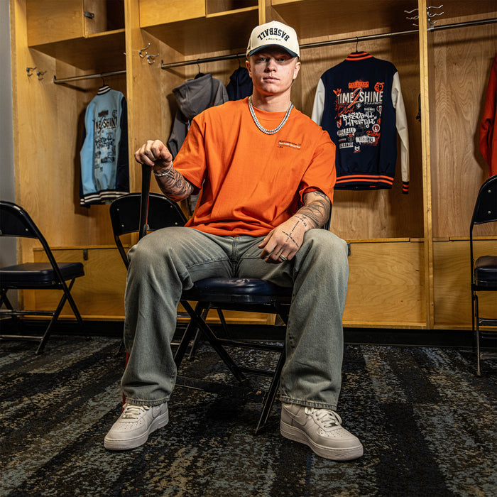 Person sitting in a locker room wearing an orange shirt and gray pants.