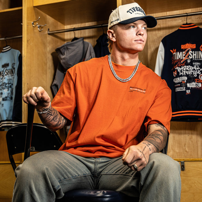 Person wearing an orange shirt and cap in a locker room with various clothing items.
