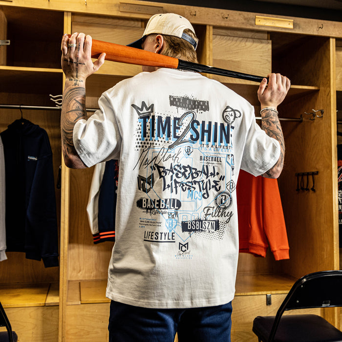 Person in a locker room wearing a white t-shirt with graphics and text, holding a baseball bat.