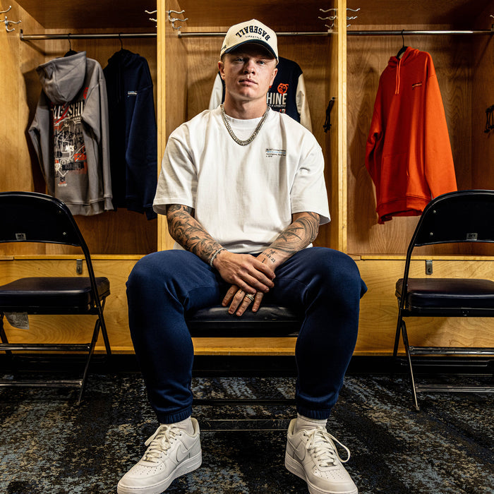 Person sitting in a locker room wearing a white t-shirt, navy pants, and white sneakers.