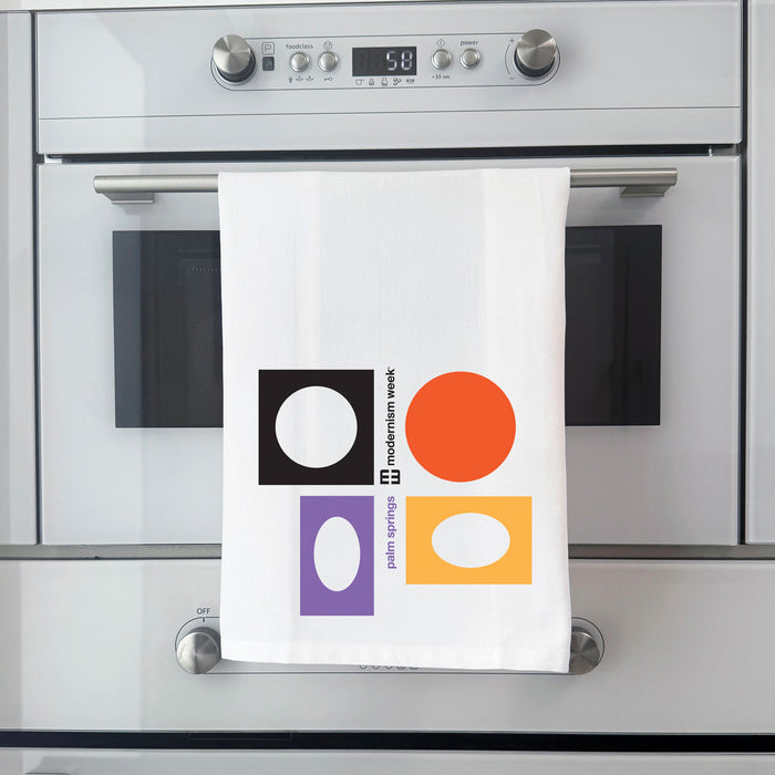 A white kitchen towel with a colorful geometric print hangs from the handle of a stainless steel built-in oven. The towel features a black square with a white circle, an orange circle, a purple square with a white oval, and a yellow-orange square with a white oval, alongside the text 'palm springs' and 'moderation week'.