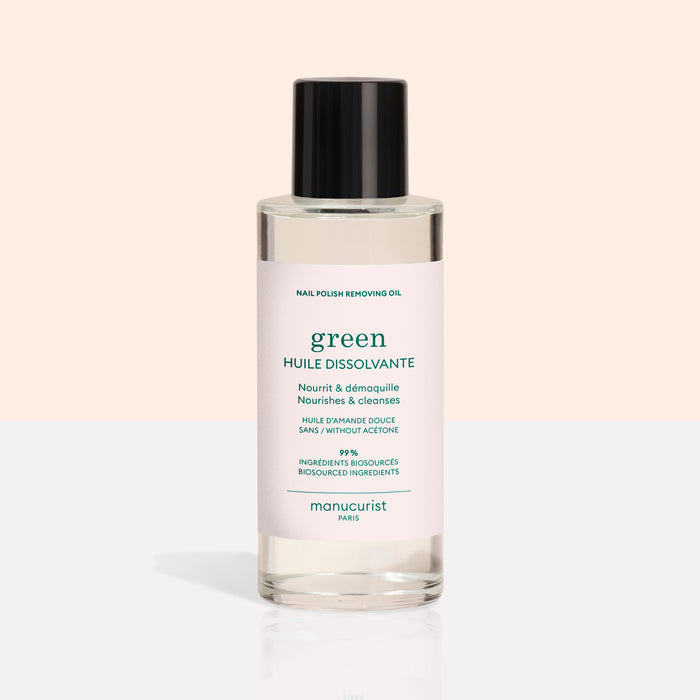 A clear bottle with a black cap contains Manucurist Nail Polish Removing Oil, a green, plant-based formula. The bottle features a light pink label with green text on a beige and white background.