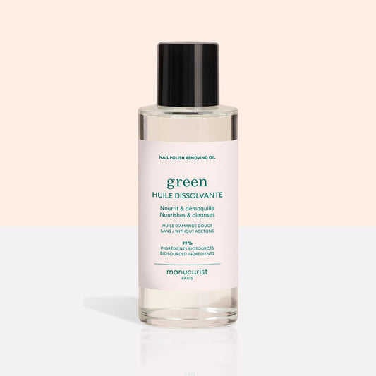 A clear bottle with a black cap contains Manucurist Nail Polish Removing Oil, a green, plant-based formula. The bottle features a light pink label with green text on a beige and white background.