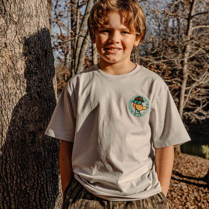 Young boy wearing a t-shirt with a duck logo, standing next to a tree in a natural setting.