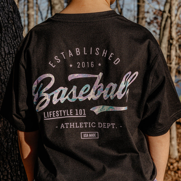 Person wearing a black t-shirt with 'Established 2016 Baseball Lifestyle 101 Athletic Dept." text in a forest setting