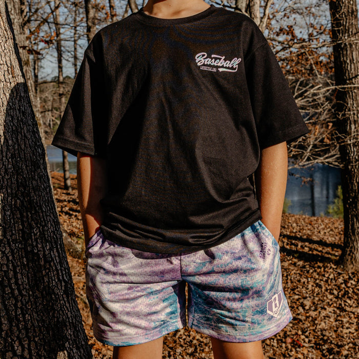 Person wearing a black t-shirt with 'Baseball Lifestyle 101' text and colorful shorts outdoors.