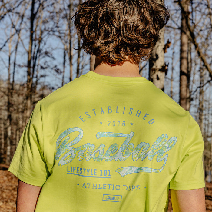 Person wearing a bright green t-shirt with 'Established 2016 Baseball Lifestyle 101 Athletic Dept." text in a forest setting