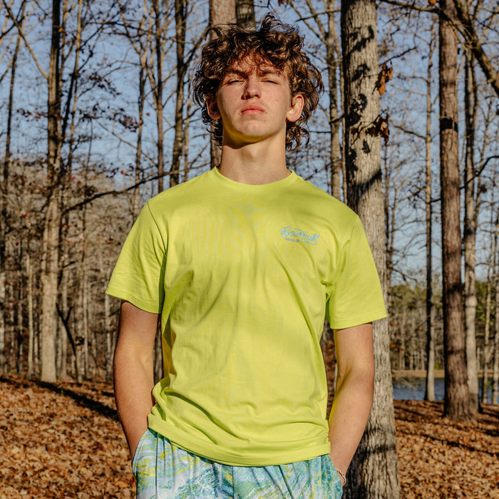 Person wearing a bright yellow t-shirt with a 'Baseball Lifestyle 101' logo in a forest setting