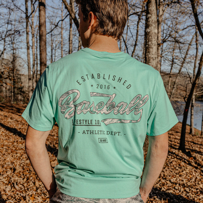 Person wearing a green t-shirt with 'Established 2016 Baseball Lifestyle 101 Athletic Dept." text in a forest setting