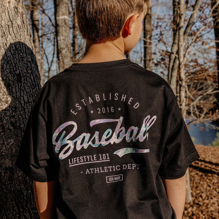 Person wearing a black t-shirt with 'Established 2016 Baseball Lifestyle 101 Athletic Dept." text in a forest setting