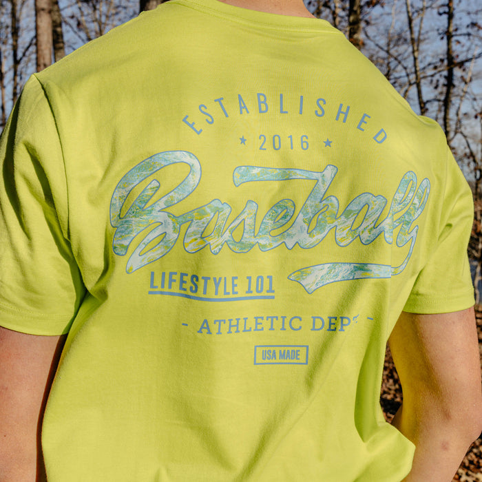 Person wearing a bright green t-shirt with 'Established 2016 Baseball Lifestyle 101 Athletic Dept." text in a forest setting