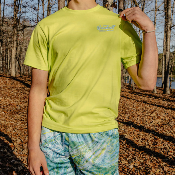Person wearing a bright yellow t-shirt with a 'Baseball Lifestyle 101' logo in a forest setting