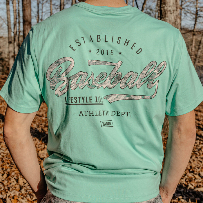 Person wearing a green t-shirt with 'Established 2016 Baseball Lifestyle 101 Athletic Dept." text in a forest setting