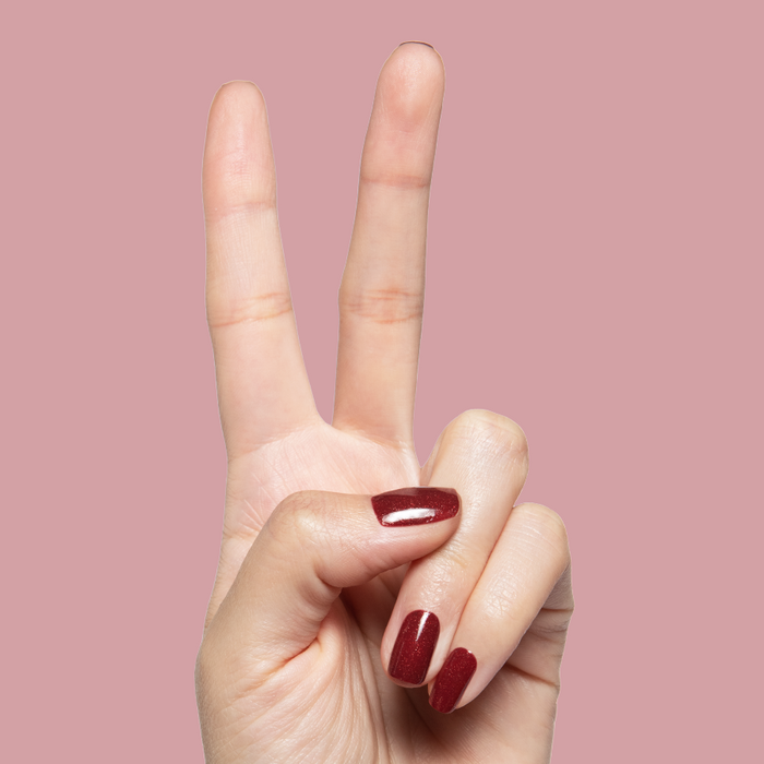 Saucy deep red shimmer quick dry nail polish shown on hand with peace sign against dusty rose background