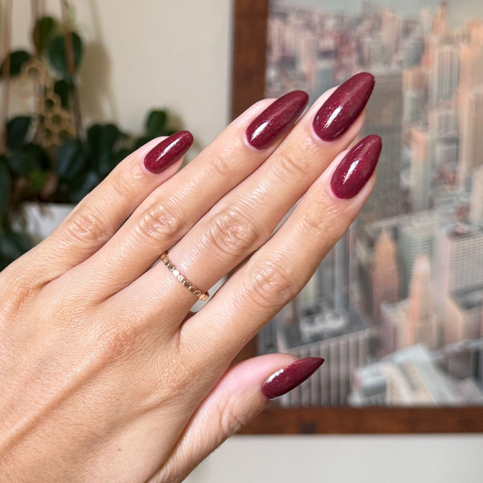 Saucy deep red shimmer quick dry nail polish shown on long almond shaped nails with cityscape background