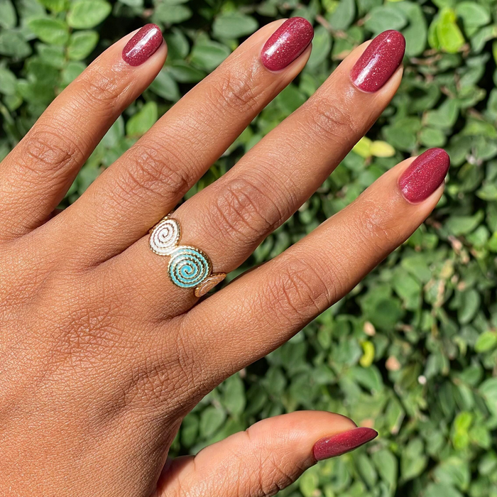 Saucy deep red microglitter nail polish on natural nails with delicate rings shown against green foliage background