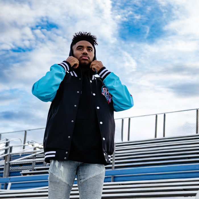 Person wearing a black and blue bomber jacket with a hood, standing against a cloudy sky.