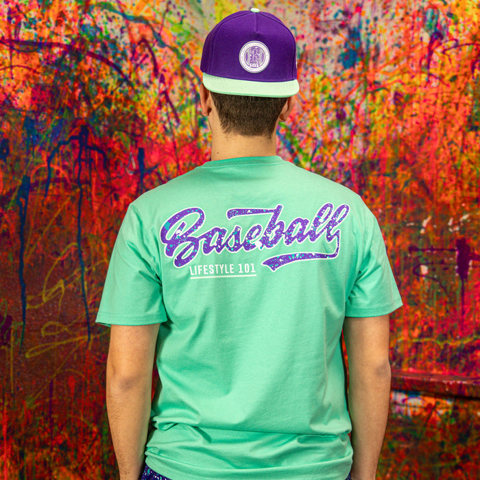 Person wearing a teal t-shirt with 'Baseball Lifestyle 101' text and a purple cap against a colorful abstract wall.