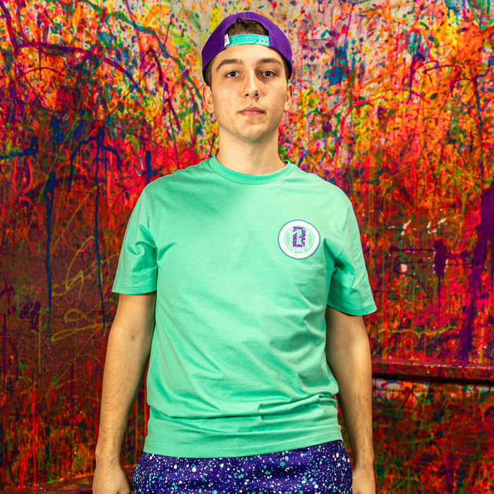 Person wearing a teal t-shirt with a logo and purple cap in front of a colorful abstract wall.