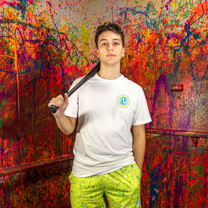 Person wearing a white t-shirt and green splatter shorts, holding a baseball bat against a colorful, abstract wall.