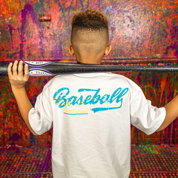 Child holding a baseball bat with 'Baseball Lifestyle 101' shirt against a colorful wall.
