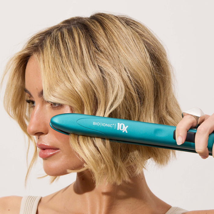 Woman using a teal hair straightener on her blonde hair against a white background